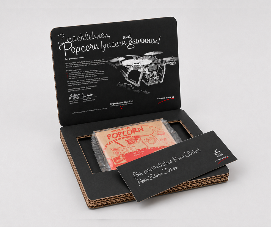 Geöffnete Versandverpackung aus schwarzem Wellkarton mit personalisiertem Kino-Give-away. Im Inneren befinden sich eine Packung Mikrowellen-Popcorn und ein personalisiertes Kino-Ticket. Der Innendeckel ist mit einer individuellen Botschaft, Illustrationen und Gewinnspielinformationen bedruckt. Die hochwertige Versandbox wird per Post zugestellt und dient als aufmerksamkeitsstarkes Direktmailing oder personalisiertes Kundengeschenk.