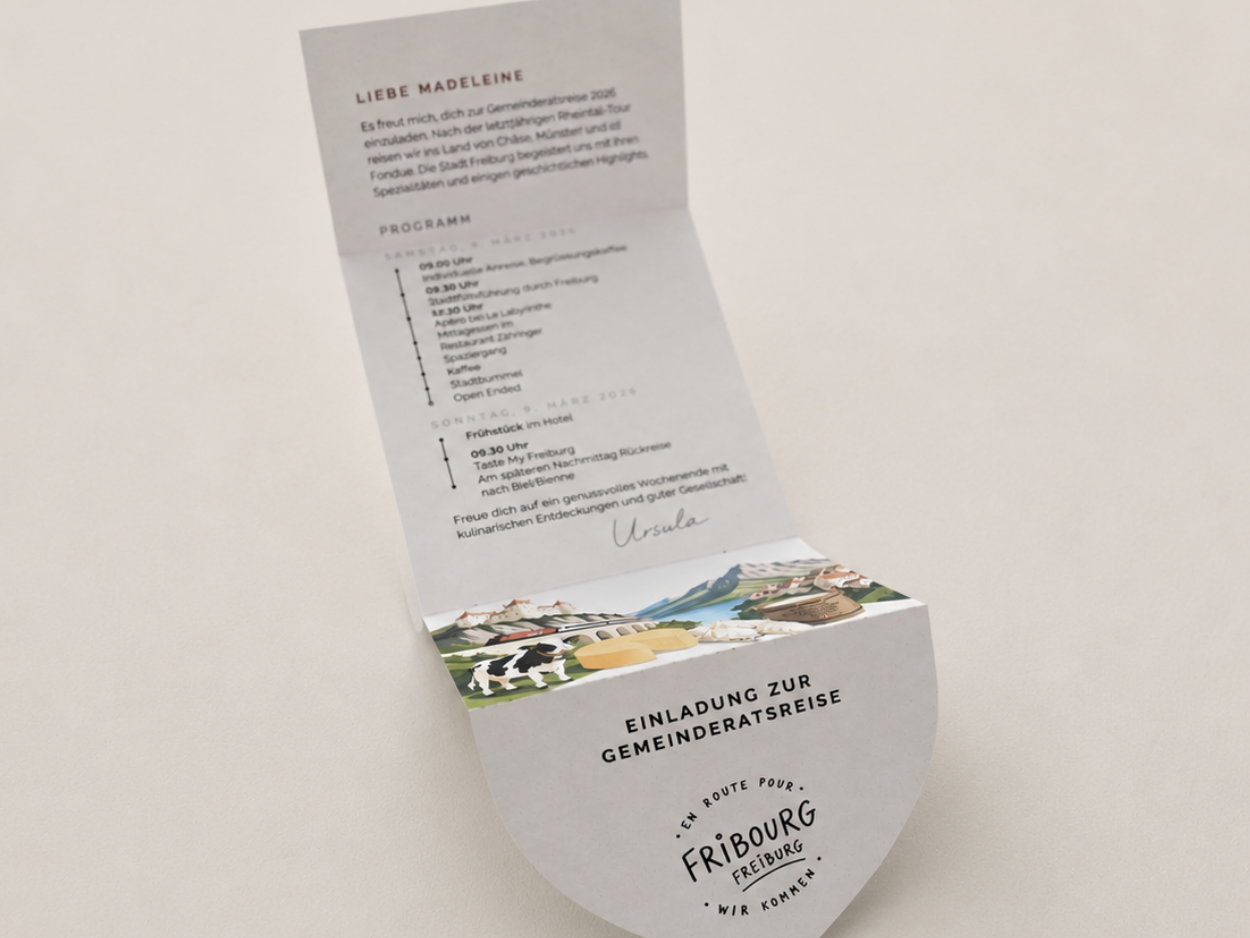 Geöffnete Einladung zur Gemeinderatsreise auf einem hellen, leicht strukturierten Hintergrund. Die aufgeklappte Karte zeigt innen ein persönliches Anschreiben sowie das Reiseprogramm, das im Hintergrund leicht unscharf erscheint. Im Vordergrund ist die halbkreisförmige Verschlussklappe mit dem Titel «Einladung zur Gemeinderatsreise» und dem Logo «En route pour Fribourg» scharf zu sehen. Eine farbige Illustration mit typischen Motiven aus dem Kanton Freiburg verbindet Innen- und Aussenseite.