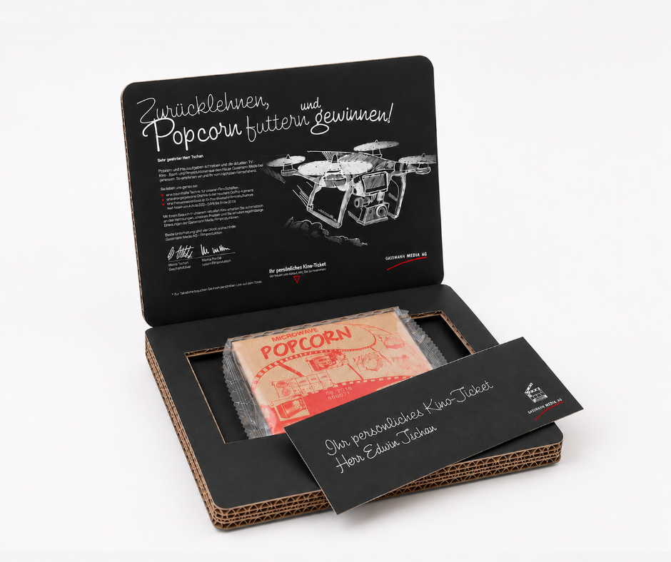 Geöffnete Versandverpackung aus schwarzem Wellkarton mit personalisiertem Kino-Give-away. Im Inneren befinden sich eine Packung Mikrowellen-Popcorn und ein personalisiertes Kino-Ticket. Der Innendeckel ist mit einer individuellen Botschaft, Illustrationen und Gewinnspielinformationen bedruckt. Die hochwertige Versandbox wird per Post zugestellt und dient als aufmerksamkeitsstarkes Direktmailing oder personalisiertes Kundengeschenk.